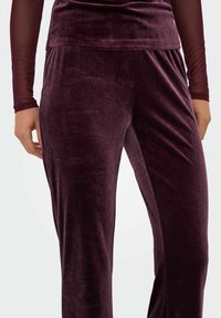 Vinrøde velourbukser med en blød tekstur, der har en bred taljebånd og en tætsiddende silhuet. Ingen synlige mønstre eller hardware-detaljer.