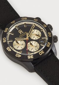 Svart chronograph klokke med gull detaljer, teksturert svart gummirem og et rundt urverk med tre sub-dialer og datovindu.