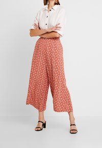 Pantalons larges à motif floral rouge avec une taille haute, en tissu léger, associés à une chemise blanche à boutons et des talons noirs.