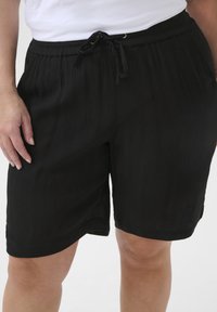 Shorts noirs plissés avec une taille élastique, cordon de serrage ajustable et poches latérales. Texture lisse, design mi-long.