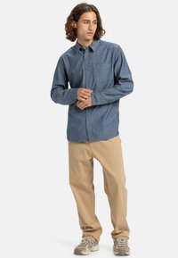 Denim button-up shirt in dark blue with a single chest pocket, paired with light beige pants, and neutral-toned sneakers.