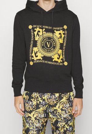 Černá mikina s zlatým barokním potiskem a nápisem „Versace Jeans Couture“ v kombinaci s kalhotami se stejným vzorem.