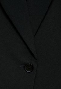 Črn blazer z ovratnikom, gladko teksturo in enim črnim gumbom z diskretnim logotipom v bližini odprtine za gumb.