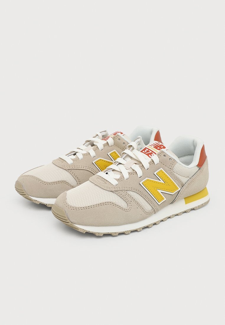 Un par de zapatillas New Balance beige con logotipos y detalles amarillos, cordones blancos y detalles naranjas en el talón, sobre un fondo claro.