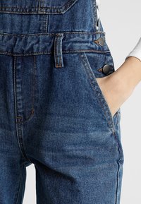 Personne portant un jean en denim bleu taille haute avec des détails de boutons, des poches avant, et une main placée dans la poche droite.