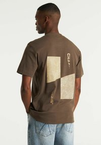 Smeđi kratki rukavi T-shirt s dva svijetlosmeđa geometrijska flastera koja sadrže uzorak linija i tekst; izrađen od mekog pamučnog materijala.