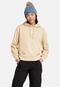 Beige hoodie med en logopatch fram, justerbara snörningar och ribbade ärmslut. Matchad med svarta cargobyxor och en blå stickad pom-pom-mössa.