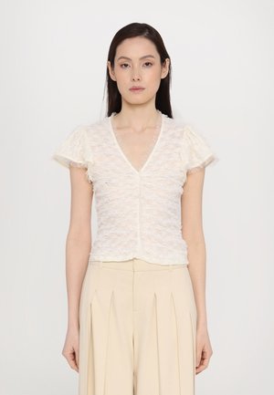 Short-sleeve, v-neck blouse in textured cream fabric. Features ruffled sleeves and a fitted silhouette, paired with high-waisted beige trousers.