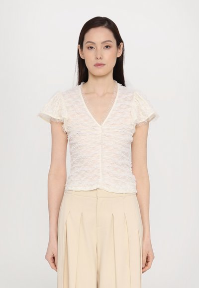 Kortetermijn, V-hals blouse in een gestructureerde crèmekleurige stof. Heeft gerimpelde mouwen en een getailleerde pasvorm, gecombineerd met hoge beige broeken.