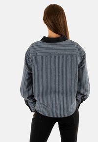 Chaqueta a rayas en azul, negro y blanco con cuello. Presenta un corte holgado y mangas abullonadas. La tela tiene una textura suave.