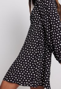 Schwarz kleid mit weißen Punkten, mit langen Ärmeln und ausgestelltem Rock. Der Stoff wirkt glatt mit einer leichten Textur.