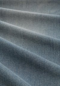 Denimstof in een lichte blauwe tint, met een gladde textuur en zichtbare weefpatronen, evenals subtiele schaduwen van gevouwen secties.