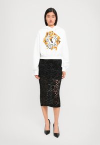 Donna che indossa un hoodie bianco con grafica dorata e una gonna nera in pizzo trasparente lunga fino al ginocchio abbinata a tacchi neri, in piedi contro uno sfondo bianco.