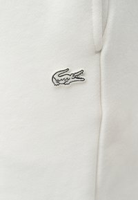 Tissu blanc avec un petit logo de crocodile brodé en noir et blanc sur la poitrine, près d'une couture et d'un cordon.