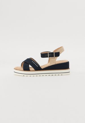 Mørkeblå sandal med to tekstilstropper, justerbar ankelrem og strukturert beige bakdel. Tykk, hvit såle med bølgete kant. Merket trykt på stroppen.