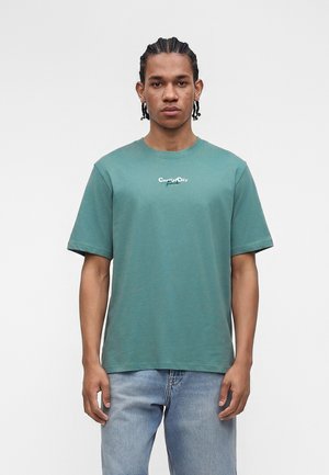 Jeune homme aux cheveux tressés portant un t-shirt turquoise « Coastal City » et un jean bleu clair, debout devant un fond blanc uni.