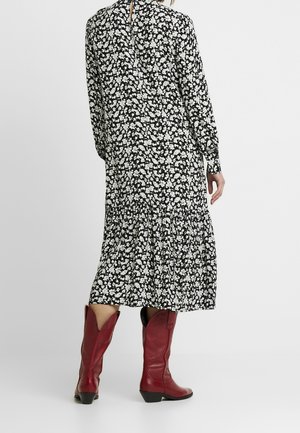 Frau trägt ein schwarz-weißes Midikleid mit Blumenmuster und rote kniehohe Lederstiefel, steht mit dem Rücken zur Kamera.