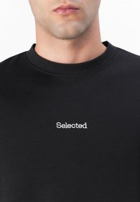 Fekete kereknyakú póló, pamutból készült, fehér hímzett "Selected" felirattal a mellkason. Simább textúra, normál szabás.