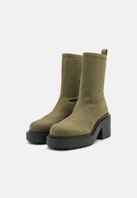 Botines verde oliva hechos de tela suave con una suela plataforma negra gruesa y una lengüeta para facilitar su colocación. Detalle de costura limpia visible.