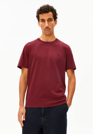 Mann mit kurzen lockigen Haaren und Schnurrbart, der ein einfarbiges burgunderfarbenes kurzärmeliges T-Shirt und dunkle Hosen trägt und vor einem weißen Hintergrund steht.