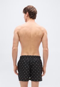 Μαύρα μαγιό σορτς με επαναλαμβανόμενο λευκό μοτίβο λογότυπου, ελαστική μέση και δύο πλαϊνές τσέπες. Ομαλή υφή με μινιμαλιστικό σχέδιο.