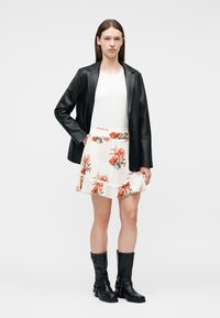 Γυναίκα που φοράει μαύρο δερμάτινο μπουφάν, λευκό τοπ, λευκή φλοράλ φούστα με κόκκινα λουλούδια και μαύρες μπότες μέσης φτέρνας, στέκεται μπροστά από μονόχρωμο φόντο.