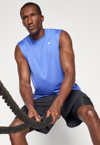 Blaues ärmelloses Sportshirt mit einem Nike-Logo, kombiniert mit schwarzen Shorts. Die Person hält ein dickes, strukturiertes Battle Rope.