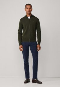 Donkergroen gebreide zip-up trui met een hoge kraag, gecombineerd met marineblauwe broek en bruine schoenen. Eenvoudig ontwerp, aansluitende silhouet, geen zichtbare patronen.