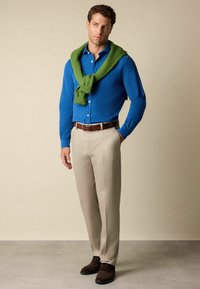 Man in een blauw overhemd met knopen, beige broek, een bruine riem en schoenen, met een groene trui om zijn schouders gegooid.