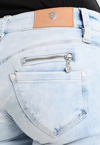 Jeans en denim bleu clair avec un patch en cuir à l'arrière, présentant une poche zippée en métal et un design de poche arrière arrondie.