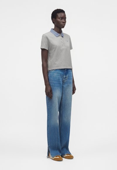 Γυναίκα φοράει γκρι πόλο μπλούζα με μπλε γιακά, φαρδιά μπλε τζιν με πλαϊνές σχισμές και ταμπά παπούτσια, στέκεται μπροστά σε απλό λευκό φόντο.