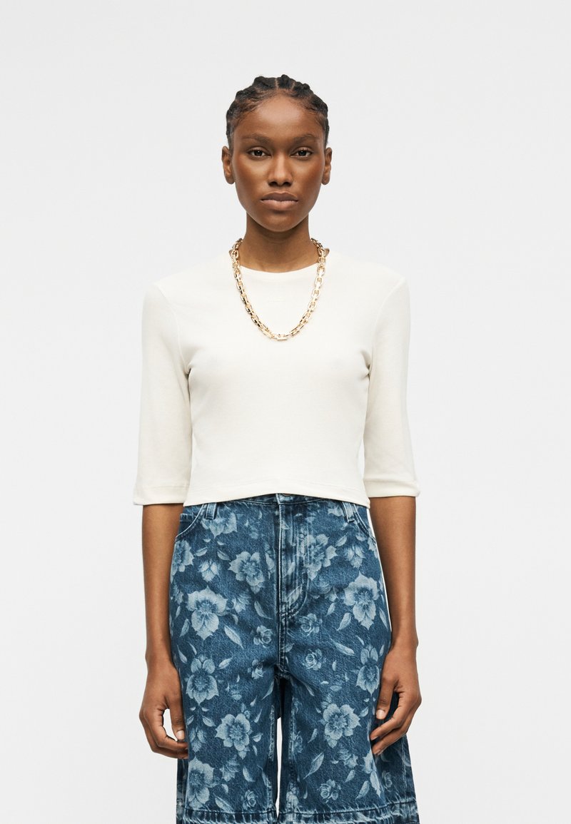 Γυναίκα με πλεξούδες που φοράει ένα λευκό cropped μακρυμάνικο τοπ, χρυσή αλυσίδα στον λαιμό και ψηλόμεσα μπλε τζιν με λουλουδάτο σχέδιο.