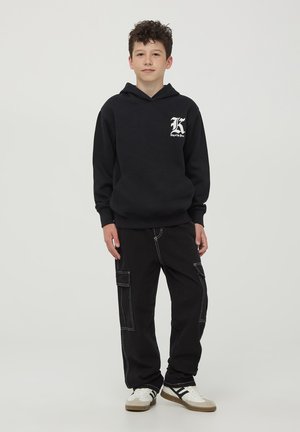 Czarna bluza z kapturem z przednią kieszenią i białym logo, w zestawie z czarnymi spodniami cargo z białymi przeszyciami i wieloma kieszeniami. Białe sneakersy.