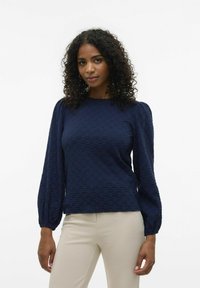 Top lungo navy a maniche lunghe con maniche a sbuffo, scollo rotondo e vestibilità rilassata, abbinato a pantaloni attillati di colore chiaro.