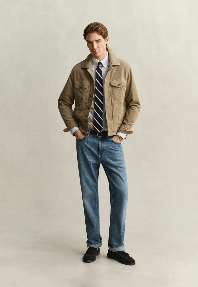 Junger Mann trägt eine beige Jacke über einem weißen Hemd mit gestreifter Krawatte, blaue Jeans und schwarze Schuhe, steht mit den Händen in den Taschen vor einfachem Hintergrund.