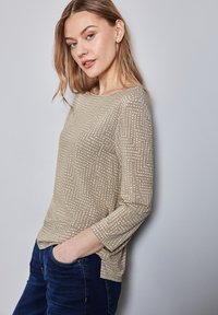 Beige Langarmoberteil mit einem strukturierten Muster aus kleinen Punkten und Linien. Weicher Stoff, runder Halsausschnitt und lockere Passform mit seitlichen Schlitzen.