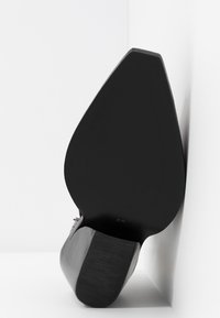 Chaussure à talon haut noire avec un bout pointu et une semelle texturée. Présente une finition brillante et un design de talon sculpté. Taille 37 affichée.