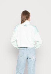 Kort vit sweatshirt med mintgröna detaljer och texturerade axlar, med hög krage och en liten Nike Air-logotyp på baksidan.