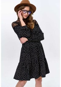 Zwarte jurk met stippen, met een gelaagde rok, kantdetails op de mouwen en een ronde halslijn, gecombineerd met een bruine hoed en grote blauwgetinte bril.