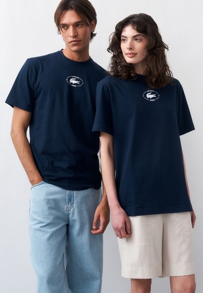 Млад мъж и жена, облечени с тъмносини тениски Lacoste Paris, застават един до друг пред едноцветен фон.