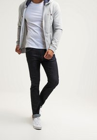 Grijze ritssluiting hoodie met een trekkoord en voork движение, gedragen over een wit T-shirt, gecombineerd met donkere jeans en witte sneakers.