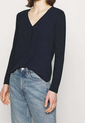 Nainen, joka pukeutunut tummansiniseen, pitkähihainen V-pääntielliseen yläosaan, joka on tungettu korkeavyötäröisiin vaaleansinisiin farkkuihin, yksinkertaisen taustan edessä.