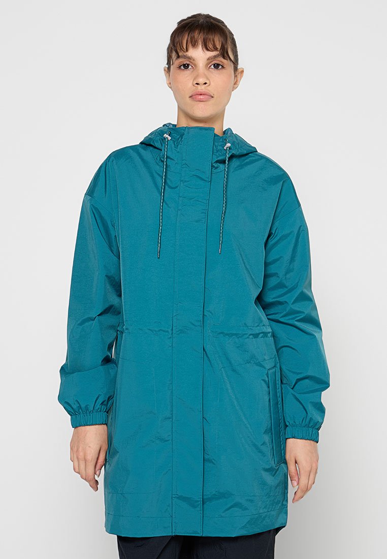 Femme portant une veste de pluie à capuche turquoise avec des poches avant et des poignets élastiques, se tenant devant un fond uni clair.