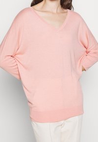 Różowy sweter z dekoltem w serek, o luźnym kroju, wykonany z miękkiej dzianiny, z opadającymi ramionami i prążkowanymi mankietami. Stylizowany z jasnymi spodniami.