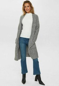 Grå stickad cardigan med öppen framsida, bärs över en vit ribbad topp, ihop med blå utsvängda jeans och svarta spetsiga ankelstövlar.