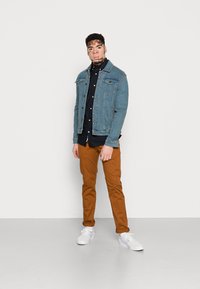 Jeansjacke in Hellblau, schwarzes Button-Up-Hemd, braune Hose und weiße Sneaker. Fokus auf lässigem, geschichtetem Outfit mit markanten Farben.
