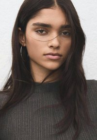 Retrato de una persona con cabello largo y oscuro, que lleva un suéter gris oscuro acanalado. Entre sus rasgos destacan los pendientes de aro y la piel suave.