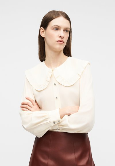 Γυναίκα με μακριά καστανά μαλλιά φοράει εκρού μπλούζα με μεγάλο βολάν στον γιακά και καφέ δερμάτινη φούστα, με σταυρωμένα τα χέρια, κοιτάζοντας πλάγια.