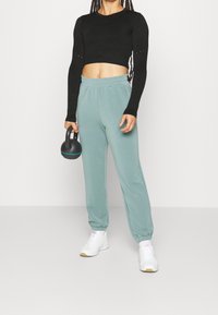 Top cropped nero a maniche lunghe, pantaloni della tuta azzurro-verdi e sneakers bianche. L'utente tiene un kettlebell nero con accenti blu.