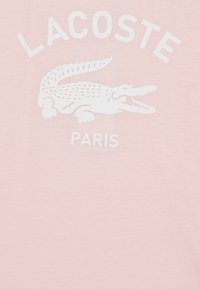 Camiseta de algodón de color rosa claro con un gráfico de cocodrilo blanco y el texto "LACOSTE PARIS" en una fuente en negrita. Textura suave y diseño casual.